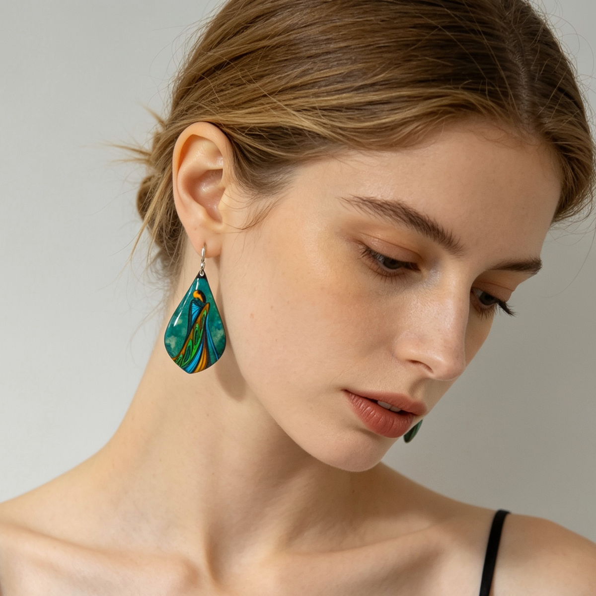 Close up of a Pair of oval earrings featuring two abstract figures surrounded by flowing blue, green and gold forms on a blond model.