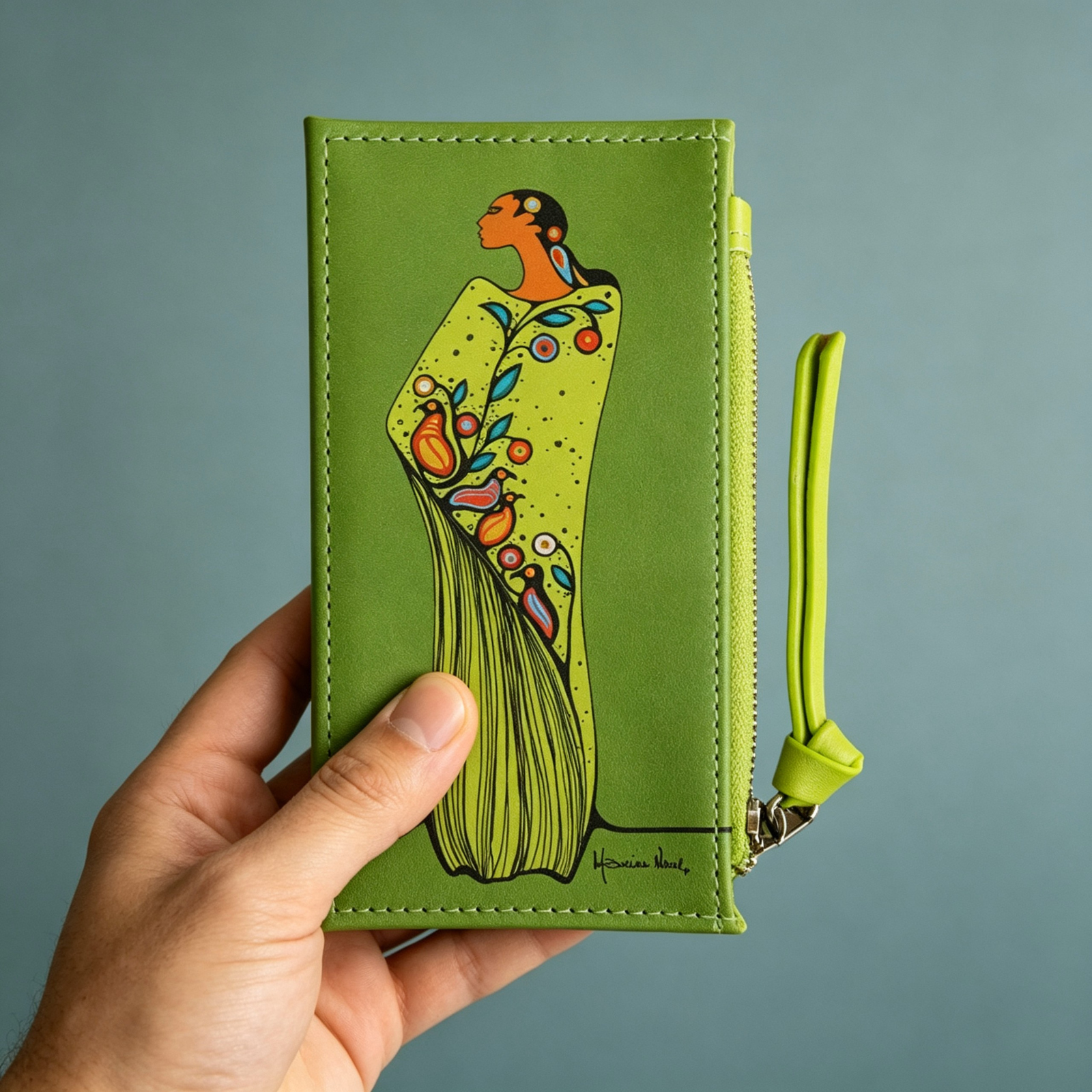 A hand holds a green leather zip-around wallet featuring lime green synthetic leather card holder featuring Maxine Noel’s Spirit of the Woodlands artwork